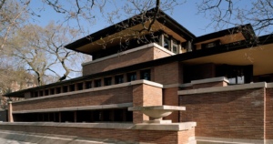 Exterior view of the Robie House showcasing its long horizontal lines, overhanging eaves, and brick and limestone structure with large windows, set against a backdrop of bare trees and a clear sky.