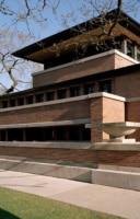 Exterior view of the Robie House showcasing its long horizontal lines, overhanging eaves, and brick and limestone structure with large windows, set against a backdrop of bare trees and a clear sky.