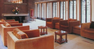 Interior view of the Robie House featuring rows of rich brown upholstered sofas arranged in a spacious room with brick walls and a large central fireplace. Art glass windows in linear designs allow natural light to enter, enhancing the warm tones of the wooden furniture and carpeted floors.