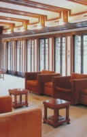 Interior view of the Robie House featuring rows of rich brown upholstered sofas arranged in a spacious room with brick walls and a large central fireplace. Art glass windows in linear designs allow natural light to enter, enhancing the warm tones of the wooden furniture and carpeted floors.