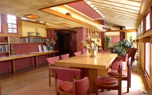 Interior of a Frank Lloyd Wright-designed residence, featuring horizontal lines, natural wood, and large tables with red chairs in an open-plan room illuminated by natural light.