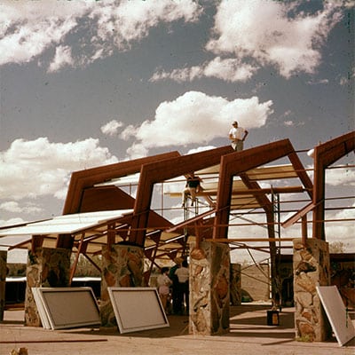 Color photograph of Taliesin West under construction, showcasing workers on the roof of a stone and wooden structure, with clear skies and desert landscape in the background.