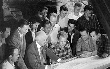 Black and white photo of Frank Lloyd Wright and a group of students gathered around a large table, examining architectural plans.