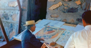 Frank Lloyd Wright, wearing a straw hat, sits at a drawing board, intently examining architectural plans in an outdoor setting with stone walls and a reflective glass window.