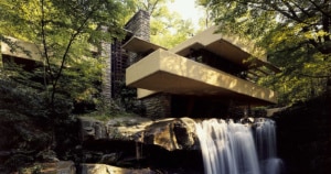 Exterior view of Fallingwater, a renowned architectural masterpiece by Frank Lloyd Wright, nestled above a waterfall amidst lush greenery. The house features multiple cantilevered balconies and terraces, extending over a natural waterfall, with a harmonious blend of concrete, stone, and glass that complements the surrounding forest.