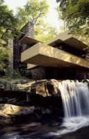 Exterior view of Fallingwater, a renowned architectural masterpiece by Frank Lloyd Wright, nestled above a waterfall amidst lush greenery. The house features multiple cantilevered balconies and terraces, extending over a natural waterfall, with a harmonious blend of concrete, stone, and glass that complements the surrounding forest.