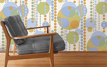A mid-century modern armchair with grey cushions in a room with a retro patterned wallpaper featuring large circles in shades of blue, green, and brown, and a polished wooden floor.