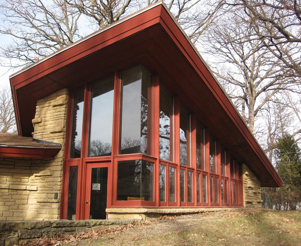 Exterior view of the Elam House, showcasing its large, vertical windows and distinctive red wooden frames, with a sloping roof and natural stone elements complementing the forested background.