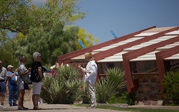 Visitors walking and talking outside a modern style building with a distinctive roof, surrounded by desert landscaping under a clear blue sky.