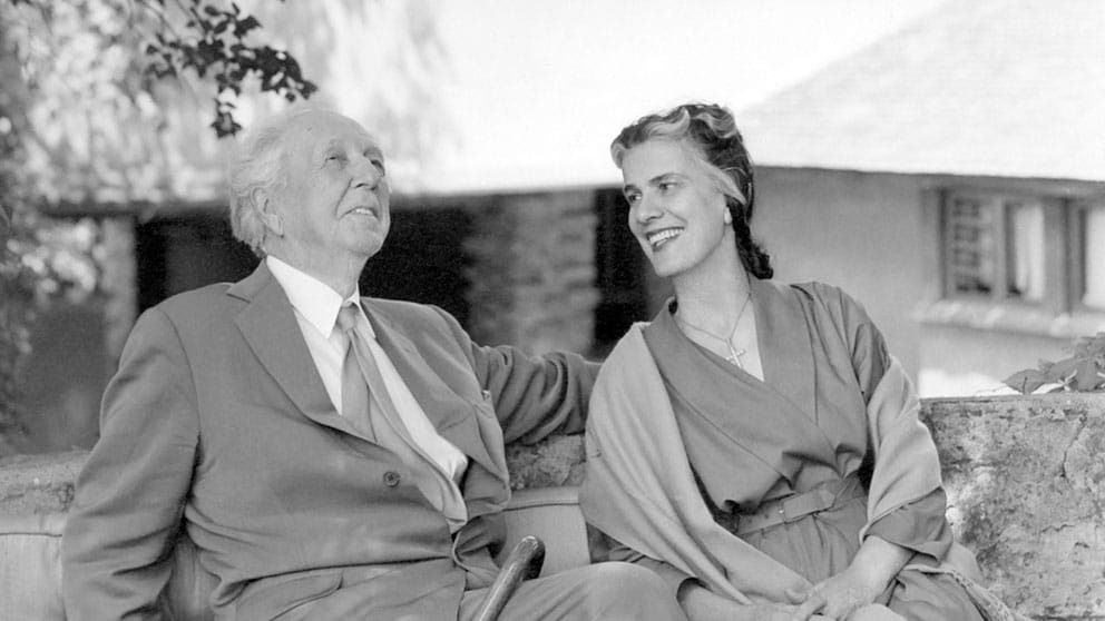 Frank Lloyd Wright and Olga Lazovich, seated next to each other, smiling, in a relaxed outdoor setting with trees and a building in the background.
