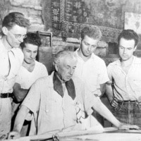 Black and white photo of Frank Lloyd Wright and a group of students gathered around a large table, examining architectural plans.