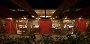 Exterior view at night of Taliesin West, showcasing warm lighting under a large sheltering structure with triangular supports, highlighting red canvas doors leading into an interior workspace visible through glass panels.