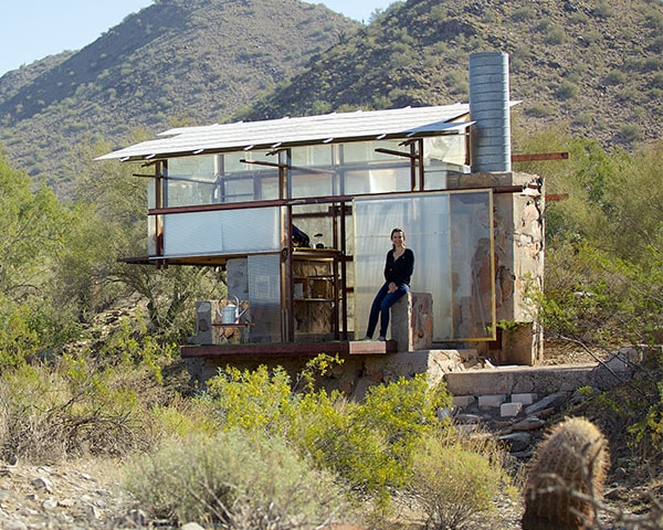 A person standing on the porch of a modern, rustic-style cabin at Taliesin West with large glass windows and metal framework, located in a desert landscape with scattered vegetation and a mountain in the background.