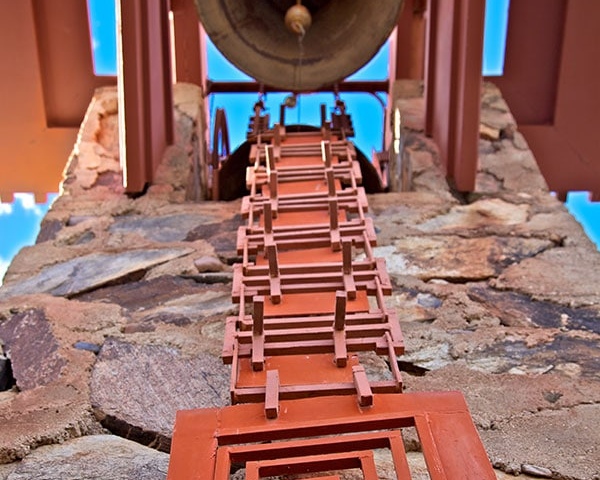 Upward view of a decorative bronze bell hanging above a geometrically arranged, red steel frame resembling a spiral staircase, set against a clear blue sky at Taliesin West.