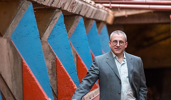 Stuart Graff standing in front of a geometrically patterned concrete structure with blue and red accents, wearing glasses and a light grey suit.