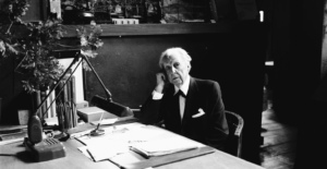 Black and white photograph of architect Frank Lloyd Wright sitting at a desk, surrounded by pencils, paper, and architectural tools, with a contemplative expression on his face.