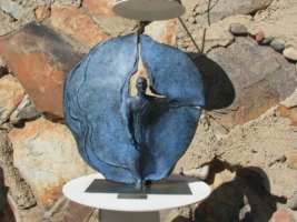 A sculpture at Taliesin West featuring a human figure emerging from a split circular blue panel, mounted on a white pedestal with a plaque, set against a natural rock background.