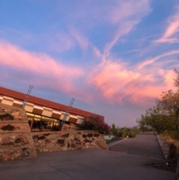 A serene evening view at Taliesin West with a pink and blue sunset sky above a building with a distinctive red-tiled roof and stone walls, flanked by native desert vegetation and a paved pathway leading to the structure.