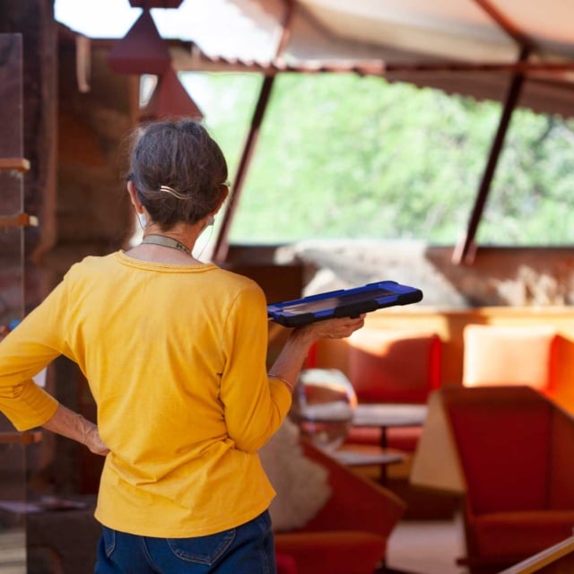 A tour guide speaking to a group of visitors at Taliesin West, the architectural school and estate designed by Frank Lloyd Wright. The room features distinctive triangular geometric shapes and is filled with natural light, with furniture consistent with Wright's design aesthetic.