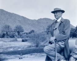 Frank Lloyd Wright wearing hat and holding cane while leaning against car with desert backdrop