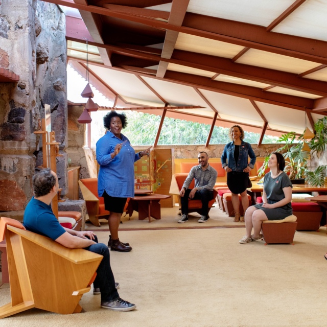 A tour guide speaking to a group of visitors at Taliesin West, the architectural school and estate designed by Frank Lloyd Wright. The room features distinctive triangular geometric shapes and is filled with natural light, with furniture consistent with Wright's design aesthetic.