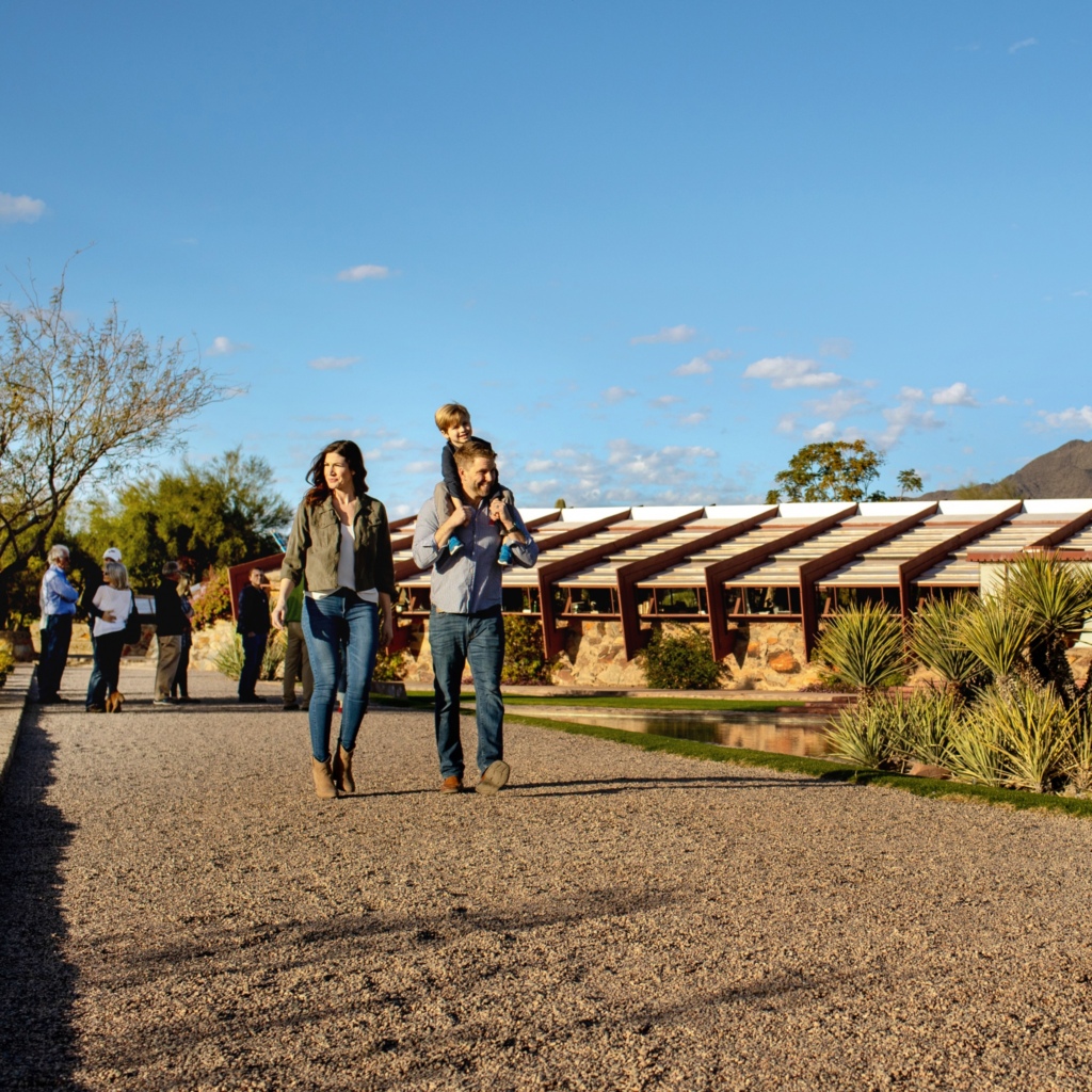 A family enjoying a sunny day at Taliesin West, with a man carrying a child on his shoulders and a woman walking beside them, with the distinctive architecture of the site in the background and other visitors milling around.