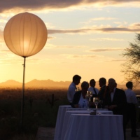 A serene sunset view at Taliesin West with guests enjoying an outdoor event under a glowing spherical balloon light, with silhouettes of distant mountains and cacti against the vibrant sky.