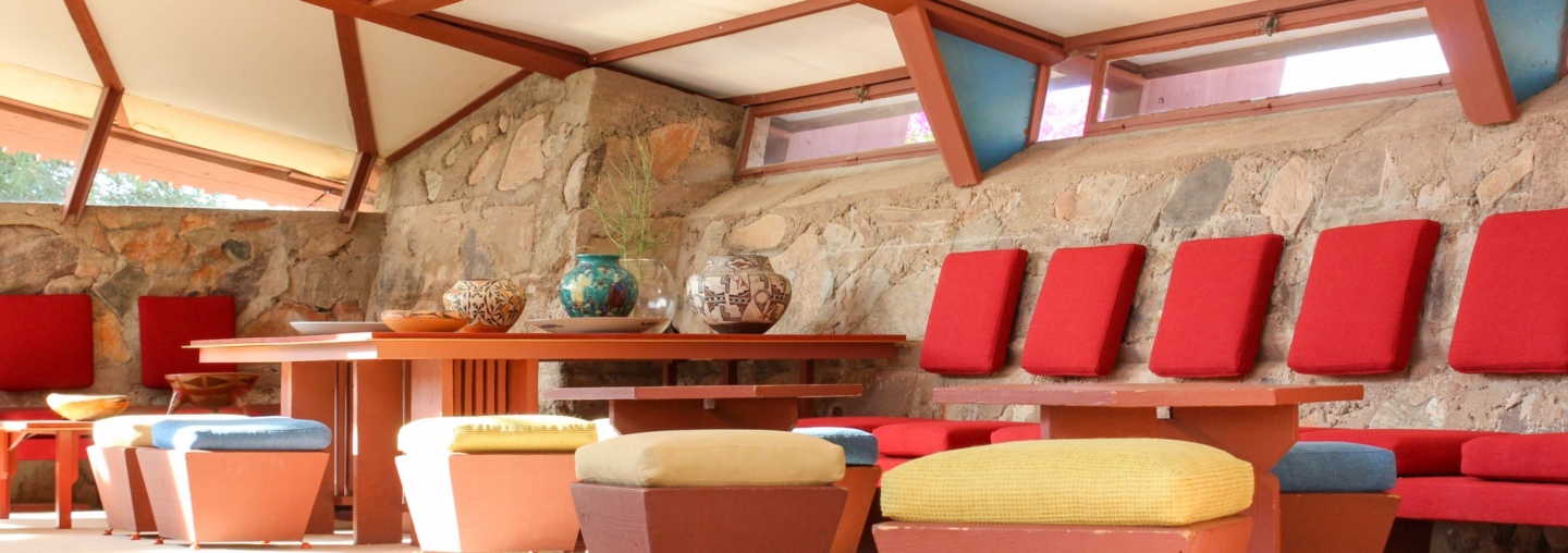 Interior view of Taliesin West showing a spacious room with stone walls and large windows under a vaulted ceiling. The room is furnished with tables and chairs in red and earth tones, and the seating includes cushioned stools and benches.