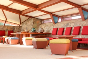Interior view of Taliesin West showing a spacious room with stone walls and large windows under a vaulted ceiling. The room is furnished with tables and chairs in red and earth tones, and the seating includes cushioned stools and benches.