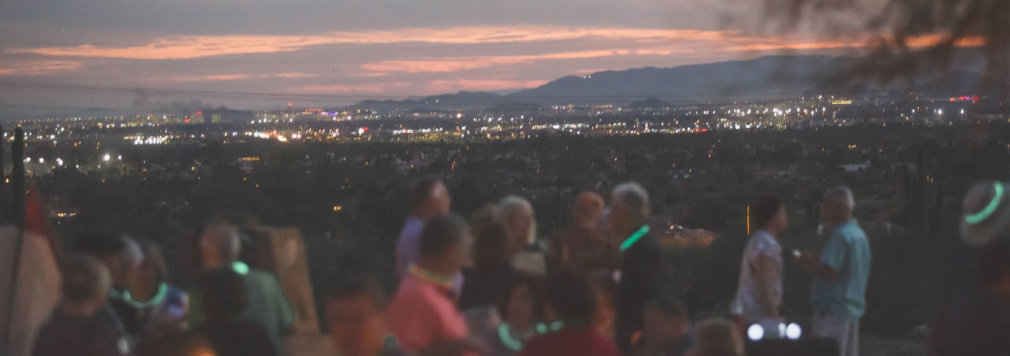 A group of people gathered at dusk, with glowing necklaces, engaging in conversation with a backdrop of a city's twinkling lights spread out beneath a twilight sky.