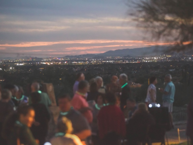 A group of people gathered at dusk, with glowing necklaces, engaging in conversation with a backdrop of a city's twinkling lights spread out beneath a twilight sky.