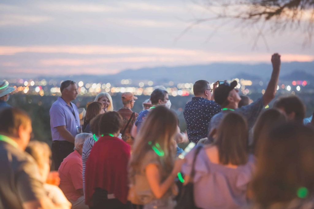 A group of people enjoying a social event outdoors at dusk, with some attendees taking photos, set against a soft-focus background featuring a sunset-lit sky and distant city lights.