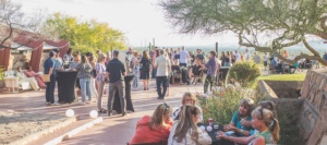 An outdoor networking event at Taliesin West at sunset, featuring people mingling and enjoying drinks, set against a backdrop of local flora, including trees and cacti, indicative of the Southwestern landscape.