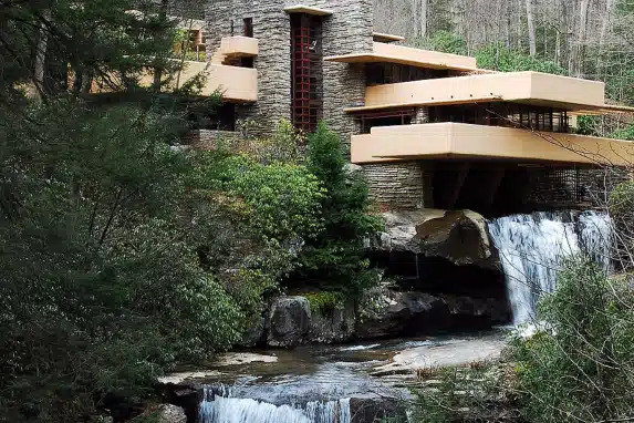 Architectural view of Fallingwater, a historic house designed by Frank Lloyd Wright, showcasing its modern cantilevered levels over a waterfall amidst lush greenery.