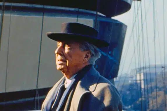 Frank Lloyd Wright in a black hat and overcoat stands outside with a clear sky behind him, looking off to the side with a contemplative expression.