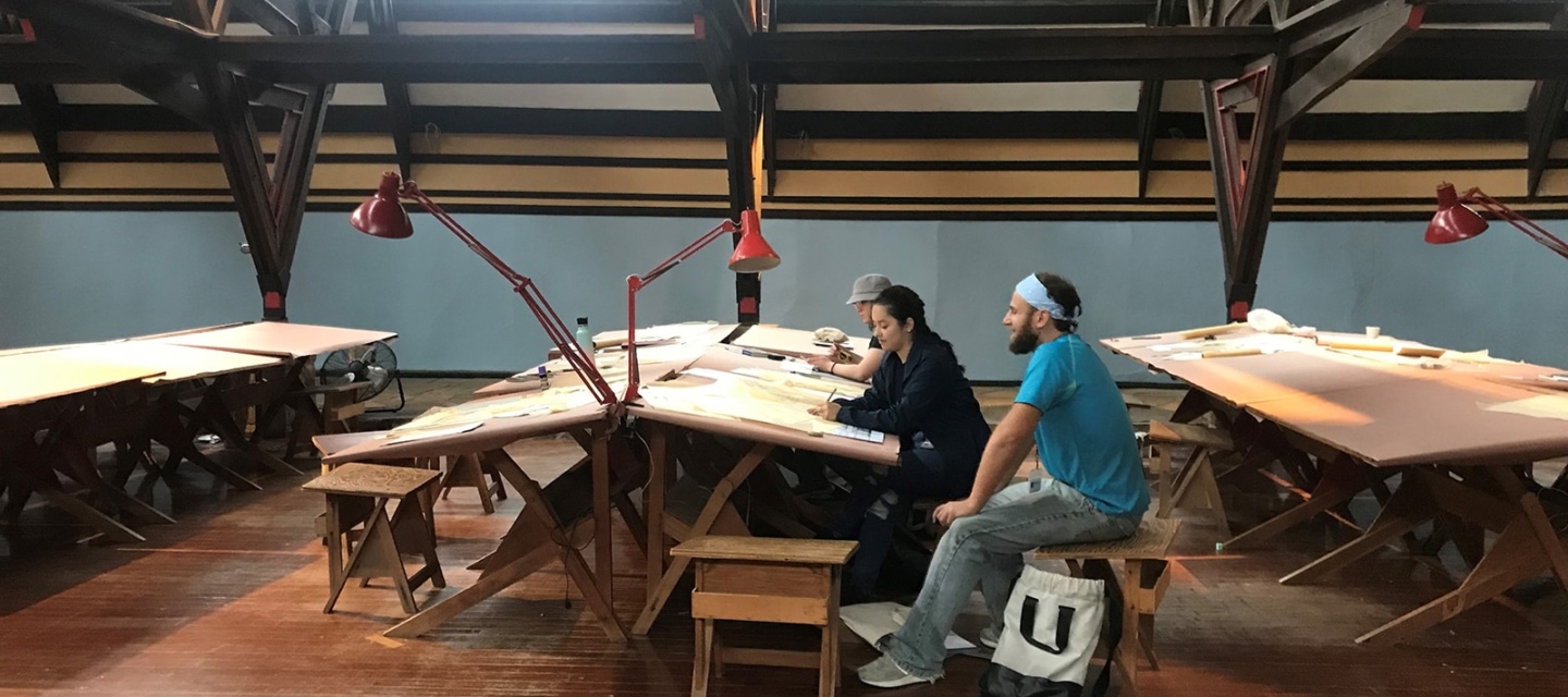 Interior of a spacious art studio with wooden floors and high ceilings featuring exposed beams. Natural light filters in through skylights. Three people are seated at long wooden tables with drawing boards, focused on their work.