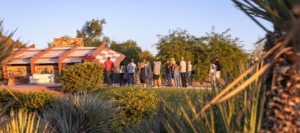A group of people on a guided outdoor tour, standing in a line and listening to a speaker, with a backdrop of unique terracotta-roofed buildings and lush desert vegetation in golden hour light.