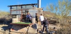 A group of people exploring an outdoor desert exhibition with a modern glass and metal structure, under a clear blue sky.