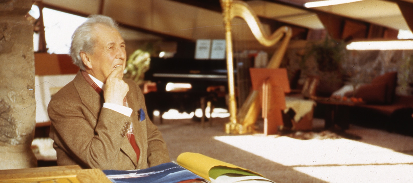 Frank Lloyd Wright working at his desk in the Garden Room at Taliesin West. Piano and harp in the background.